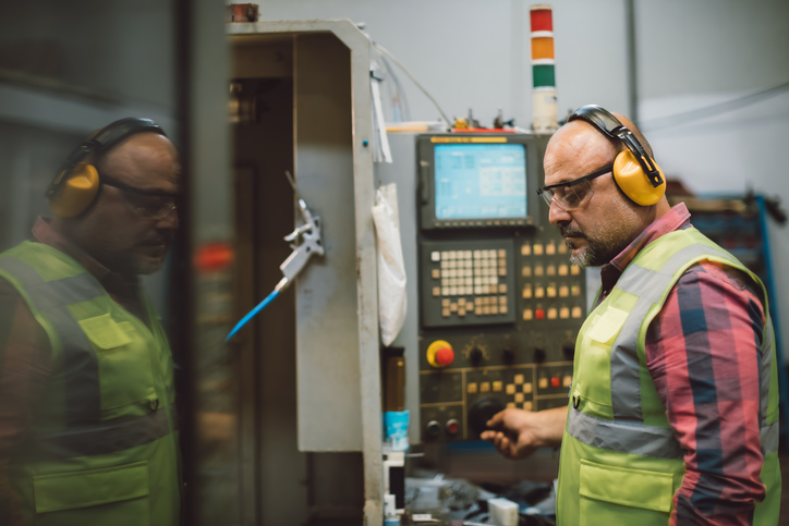 Middle aged male industrial worker wearing high visibility vest and safety glasses operating CNC manufacturing machine in a modern factory environment with automated production systems, heavy machinery, digital control panels, smart manufacturing technology and industrial workflow in a large production facility