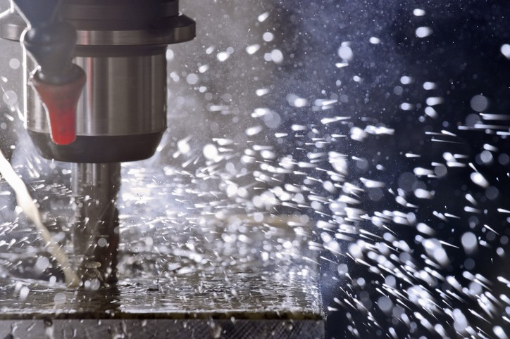 A strongly back-lit photograph of a precision CNC machining tool in operation. Purposely lit and styled to capture the extreme amount of coolant spray that fills these closed manufacturing environments.