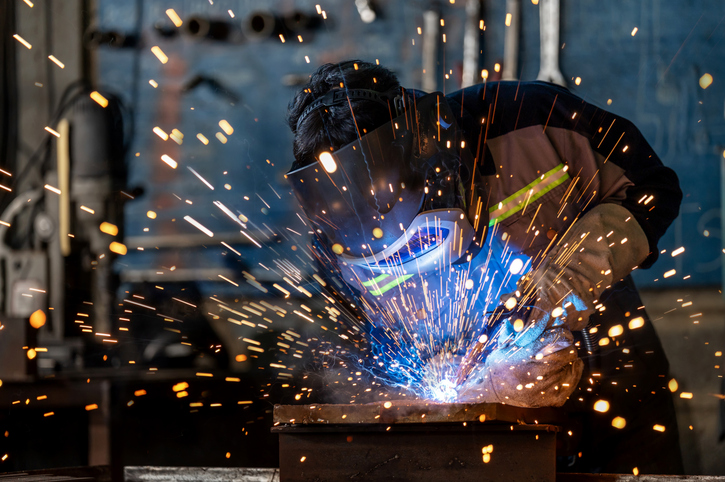 A professional welder wearing full protective gear works with steady hands and focus, welding a metal component. The vibrant sparks create a beautiful bokeh that contrasts the intense environment with artistic energy, showcasing skill and dedication.