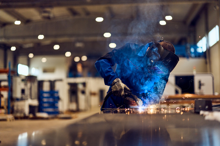 Worker welding metal in a factory workshop, surrounded by sparks and blue smoke.
