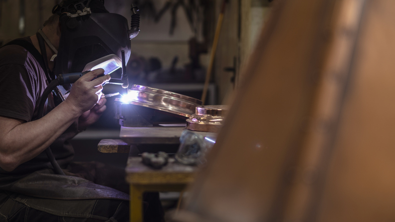 The welder wearing the welding mask working on the brass details with the electrical welding, with the blue flame and smoke around the welding point.